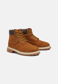 Stivali alla caviglia in pelle marrone con lacci gialli, colletto imbottito e suola in gomma bianca con un motivo testurizzato. Etichetta con il logo Timberland attaccata.