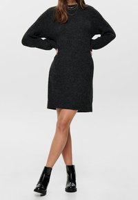 Robe-pull noire à coupe décontractée, texture côtelée et manches longues, associée à des bottines noires. Finition lisse et longueur jusqu'aux genoux.