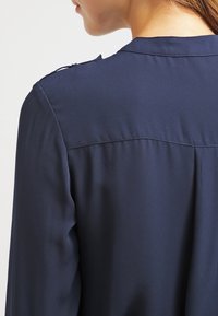 Blusa blu navy con una texture liscia, dotata di colletto a camicia e dettagli sottili sulle spalle. Il retro presenta delle pieghe per una maggiore struttura.