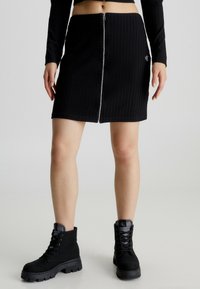 Falda negra de canalé con cremallera en el frente y un pequeño detalle de logo. Combinada con botas negras hasta el tobillo con suela gruesa y diseño de cordones.
