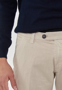 Pantaloni di cotone beige con chiusura a bottone, dotati di tasche laterali e una texture di tessuto liscia. Abbinati a un maglione blu scuro.