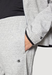 Γκρι φούτερ με φερμουάρ και πλαϊνές τσέπες, σετάρισμένο με αντίστοιχο γκρι παντελόνι. Δ ιαθέτει μαύρη λογότυπο λεπτομέρεια και αντίθετη μαύρη επένδυση στα μανίκια.