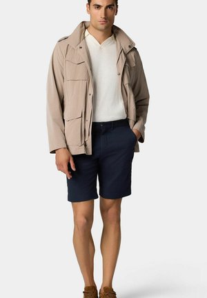 Veste légère beige avec plusieurs poches, portée par-dessus une chemise blanche à col en V. Associée à un short marine et des mocassins marron.