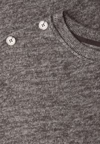 Grå stickad tröja med rund halsringning och två vita knappar på axeln. Mjuk textur med ett diskret mönster.