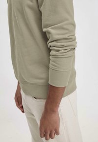 Olivgrüner Sweatshirt mit gerippten Bündchen und Saum, aus weichem Stoff gefertigt, neben hellen Hosen präsentiert. Arm entspannt an der Seite positioniert.