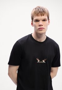 Svart bomulls T-skjorte med brodert design av to fugler og ordet "HUGO" i midten, med en klassisk rund hals.