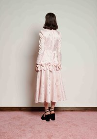 Kobieta stoi twarzą do ściany, ubrana w jasnoróżową kwiecistą sukienkę midi z kokardami w talii oraz czarne sandały na wysokim obcasie, na różowym dywanie.