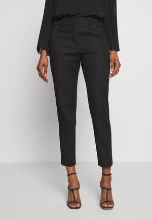 Femme portant un pantalon noir slim longueur cheville, un haut noir plissé à manches longues et des sandales noires à talons hauts avec des lanières, debout devant un fond uni.