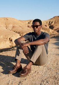 Homme portant des lunettes de soleil, une chemise sombre, un pantalon à motifs et des chaussures marron, assis sur un sol sec et rocheux avec des collines désertiques en arrière-plan.