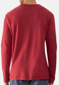 Man in een effen rode lange mouwen shirt en marineblauwe gebloemde broek, gezien van achteren tegen een witte achtergrond.