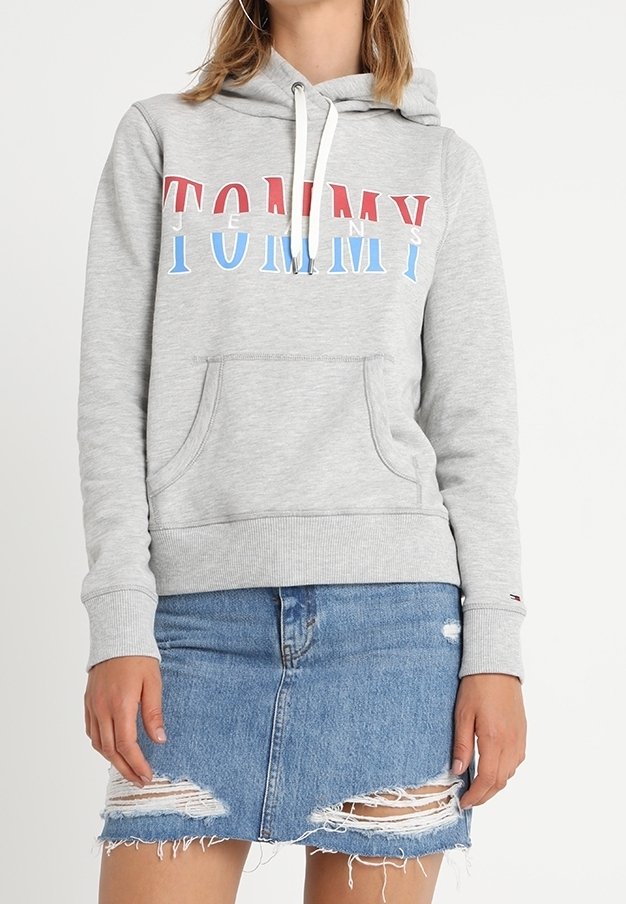 Femme portant un sweat à capuche gris clair avec "TOMMY JEANS" en lettres rouges, blanches et bleues et une mini-jupe en jean bleu déchirée.