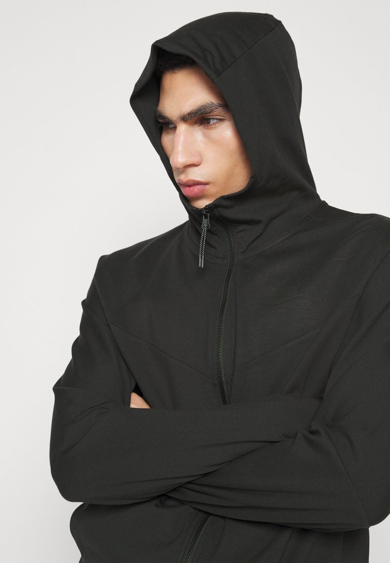 Felpa nera con zip, colletto alto e cappuccio regolabile. Presenta un logo discreto, linee eleganti e una texture di tessuto liscia.