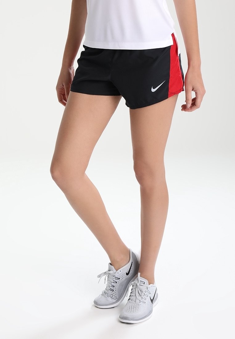 Personne portant un short de sport Nike noir et rouge, un t-shirt blanc, et des chaussures de course Nike blanches, debout sur un fond blanc.