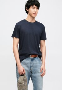 Tumši zila īsroku T-krekls no mīksta auduma, valkāts ar gaiši zilām džinsu biksēm un brūnu jostu, turot pelēlu beisbola cepuri.