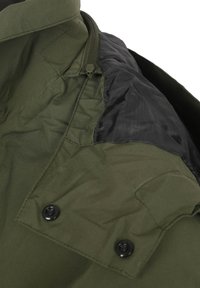 Olivengroene jas met een zwarte voering in de kraag. Beschikt over een ritssluiting, een gemêleerde stof en twee zwarte knopen aan de voorkant.