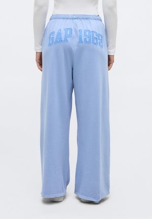 Osoba nosící světle modré široké tepláky s výšivkou "GAP 1969" na zadním pásu, bílé tričko a bílé boty.