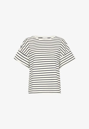 Nainen seisoo sisätiloissa päällään valkoinen ja musta raidallinen lyhythihainen paita, vaaleansiniset farkut ja beiget balleriinat mustilla pilkuilla.