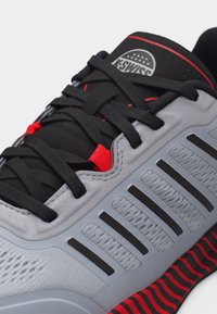 Grau-schwarzer Sneaker mit schwarzen Schnürsenkeln, roten Akzenten und einer strukturierten Sohle mit rot-schwarz gestreiftem Muster.