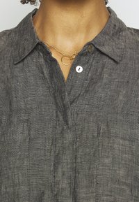 Šedá lněná košile s límečkem, má knoflíkový výstřih a přední kapsy. Jemný zlatý náhrdelník s prsteny zdobí výstřih.