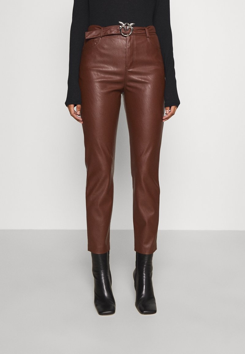 Femme portant un pantalon en cuir marron taille haute avec une ceinture à boucle argentée, un haut noir à manches longues et des bottines noires, debout devant un fond uni.
