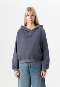 Grå cropped hoodie med snørre, der har et logo på brystet og rummelige ærmer. Båret med lyseblå vide jeans.