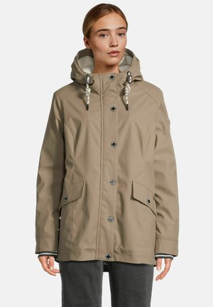 Vrouw in een beige hoodie-jack met knoopsluitingen en voorflapzakken, staand tegen een effen achtergrond.