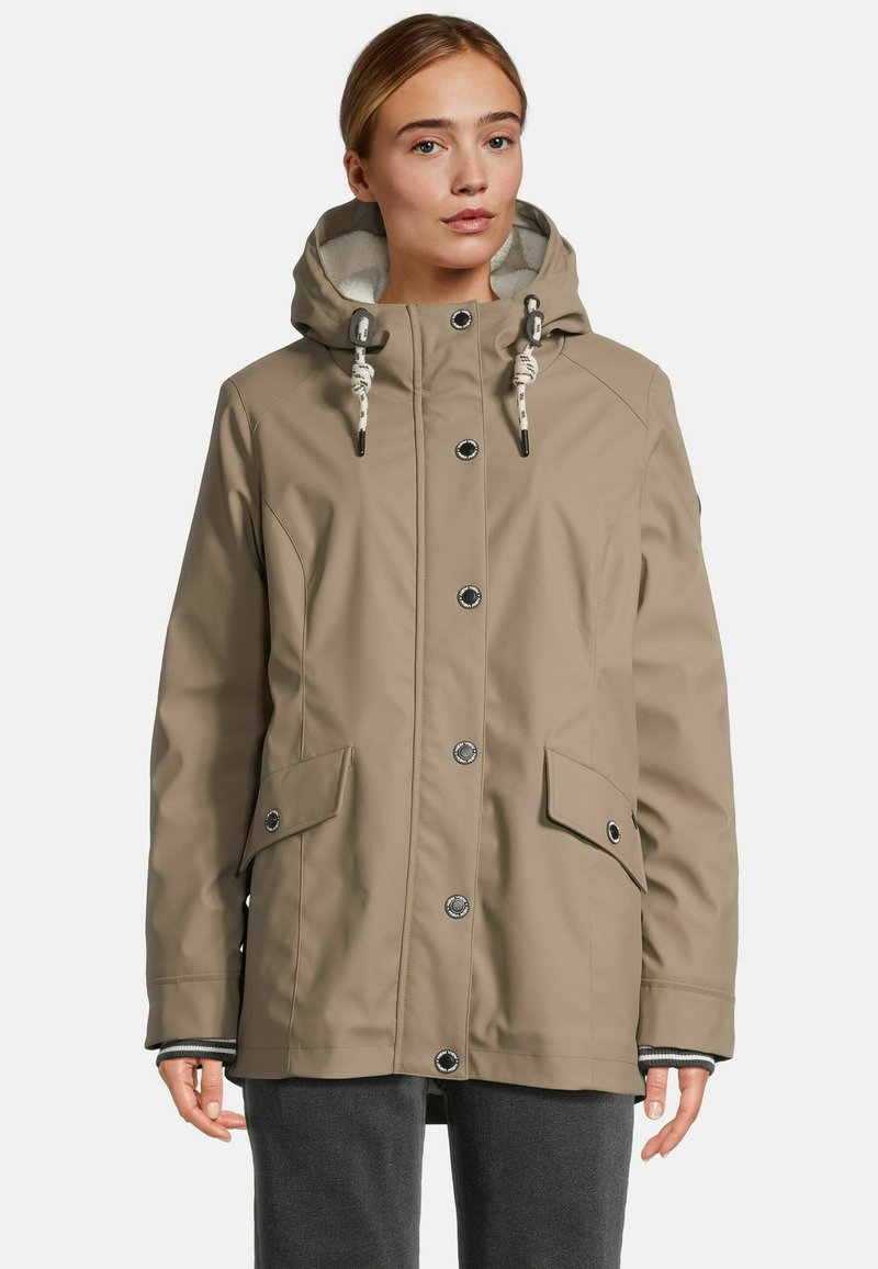 Femme portant une veste à capuche beige avec des fermetures à bouton et des poches à rabat à l'avant, se tenant devant un fond uni.