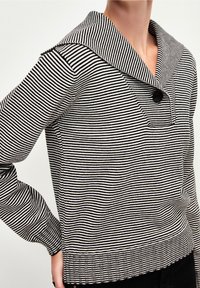 Pull à rayures noir et blanc avec un grand col, des boutons décoratifs et un tissu texturé. Manches longues et finitions à bords côtes.