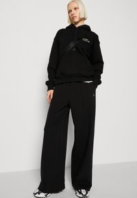 Felpa nera con cappuccio e tasca frontale, abbinata a pantaloni neri a gamba larga. Accento notevole del logo verde sulla felpa e scarpe nere con motivo.