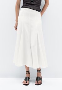 Jupe évasée blanche avec une texture lisse, arrivant au genou et présentant des plis discrets. Associée à des sandales noires à lanières.