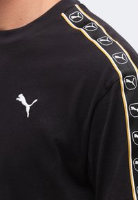 Μαύρο πουκάμισο με λευκό λογότυπο Puma στο στήθος και επαναλαμβανόμενα λογότυπα Puma σε μαύρη λωρίδα με κίτρινες άκρες κατά μήκος της ραφής του ώμου.