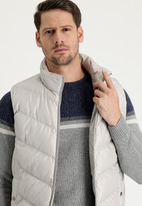 Chaleco acolchado de color gris claro con cuello alto y cremallera, llevado sobre un suéter texturizado de color azul oscuro que presenta rayas blancas y grises.