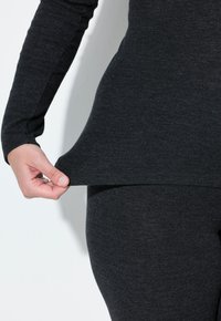Schwarzes Langarmoberteil mit abgerundetem Saum. Der Stoff wirkt weich und strukturiert. Eine Hand zieht am unteren Rand, wodurch die eng anliegenden schwarzen Leggings darunter sichtbar werden.