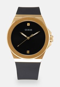 Reloj con esfera negra y caja en tono dorado, manecillas y índices dorados. Presenta una correa de silicona negra y una corona en el lado.