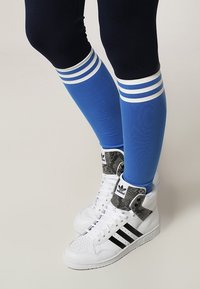 Bílé vysoké tenisky s černými pruhy a šedými akcenty, v kombinaci s modrými podkolenkami se bílými pruhy a černými legínami.