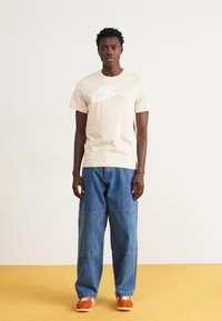 Camiseta beige de Nike con gran logo blanco, combinada con vaqueros de mezclilla azules holgados y zapatillas naranjas. Fondo liso, pose de pie.