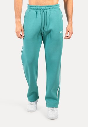 Türkise Jogginghose mit einem Kordelzugbund, zwei weißen Seitennähten und einem Logo am linken Oberschenkel, kombiniert mit weißen Turnschuhen.