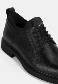 Chaussure en cuir noir avec une surface texturée, bout arrondi et design à lacets. Dotée d'une semelle en caoutchouc robuste et d'une boucle à l'arrière pour un enfilage facile.
