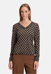 Maglietta nera a maniche lunghe con scollo a V e motivo geometrico beige. Presenta un colletto a coste e un tessuto morbido e leggero. Indossata con pantaloni marroni.