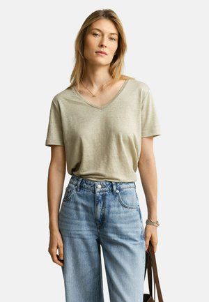 Blonde Frau trägt ein hellbeiges V-Ausschnitt-T-Shirt, das in eine hoch taillierte hellblaue Jeans gesteckt ist, sie hält den Riemen einer braunen Tasche in der rechten Hand.