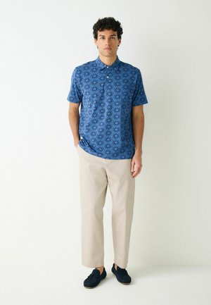 Bărbat stând cu mâinile în buzunare, purtând un tricou polo albastru cu model, pantaloni bej și pantofi negri pe fundal alb.