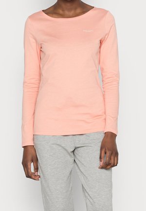 Persona vestida con una camiseta de manga larga rosa claro y pantalones gris claro, de pie frente a un fondo liso.