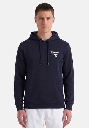 Uomo che indossa una felpa blu navy con tasca frontale e logo bianco sul petto, abbinata a pantaloni bianchi, in piedi su uno sfondo semplice.