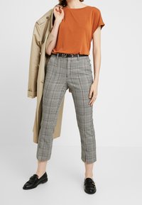 Brown short-sleeve top, gray plaid trousers with side slits, black belt, and black loafers. Beige trench coat draped on one shoulder.