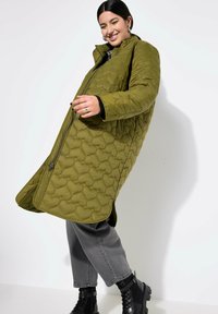 Cappotto trapuntato verde oliva con colletto alto, decorato con zip frontale e spacchi laterali. Indossato sopra pantaloni grigi e stivali neri alla caviglia.