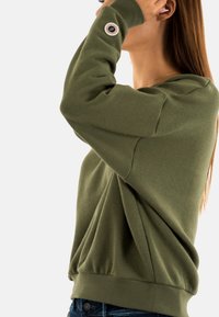 Sudadera verde oliva con un corte relajado, textura suave y puños de canalé. Detalle notable: un acento de logo circular en la manga.
