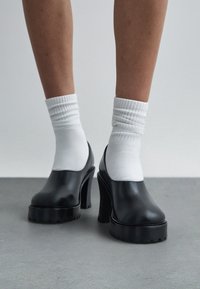 Άτομο που φορά μαύρα χοντρά πλατφόρμα τακούνια σε συνδυασμό με λευκές κάλτσες, στέκεται σε γκρι πάτωμα μπροστά σε απλό φόντο.
