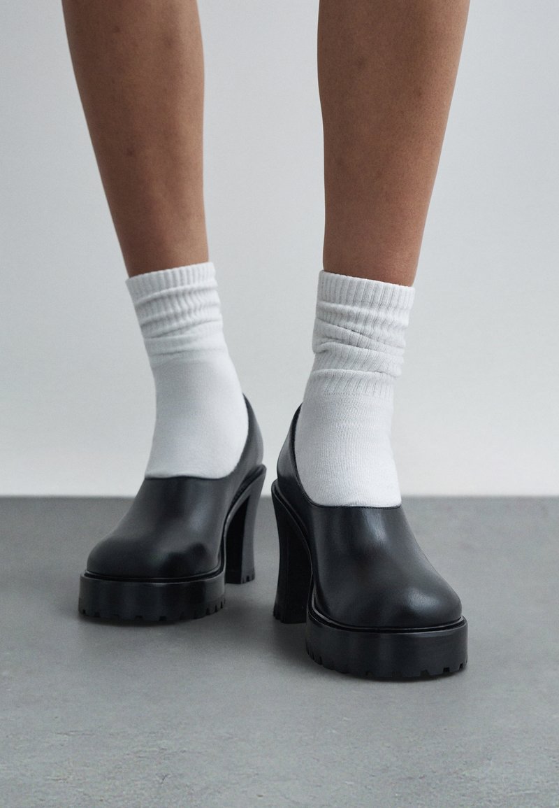 Άτομο που φορά μαύρα χοντρά πλατφόρμα τακούνια σε συνδυασμό με λευκές κάλτσες, στέκεται σε γκρι πάτωμα μπροστά σε απλό φόντο.