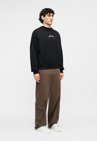 Νεαρός που στέκεται, φοράει μαύρο φούτερ με μικρό λευκό λογότυπο, καφέ παντελόνι cargo και λευκά αθλητικά παπούτσια, σε απλό λευκό φόντο.