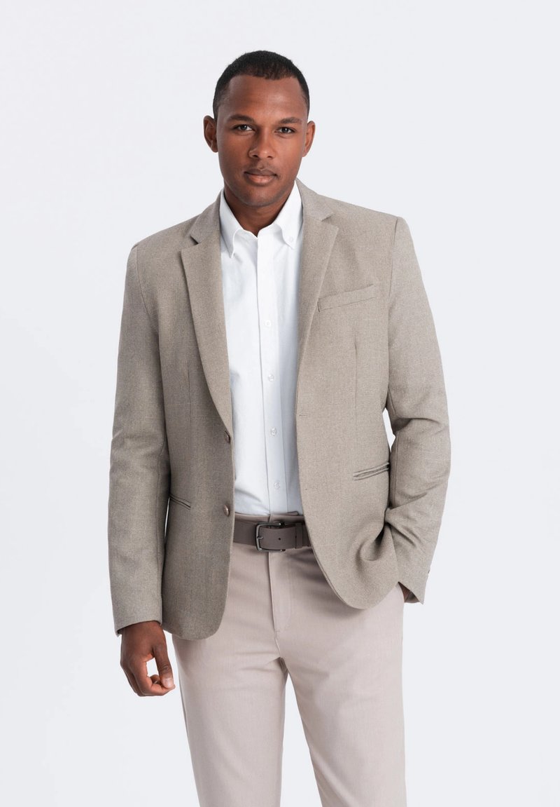 Blazer marron clair avec une coupe ajustée, fermeture à un bouton, revers crantés et poches latérales, associé à une chemise blanche et un pantalon clair.